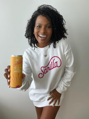 Faith reaching out a can of our lemon spritzer and smiling in a cozy white Sarilla sweatshirt, styled for a relaxed fit. The image captures the soft texture and versatile design, perfect for everyday comfort